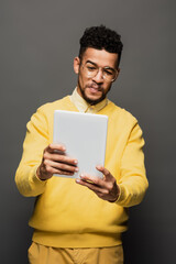 displeased african american man in glasses using digital tablet isolated on grey.