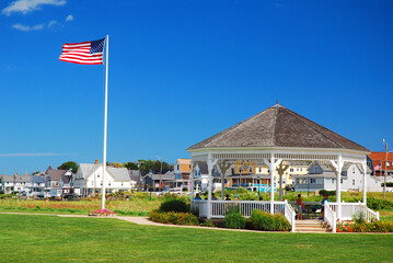 An American flag flies in a town square with a gazebo on a sunny summer day