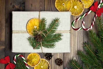 The Christmas gift is wrapped in linen cloth. A gift, spruce branches, orange slices, lollipops on a wooden table. The concept of Christmas and New Year holidays in the style of zero waste. Top view.