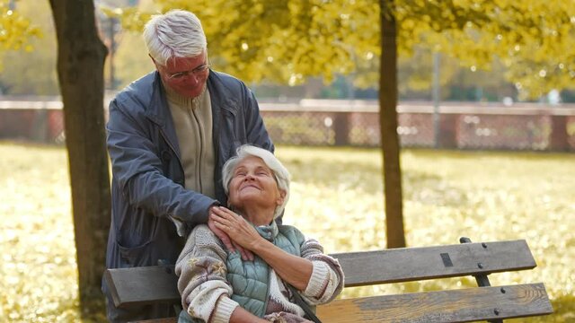 An elderly couple in the park. Man placing his hand over the shoulder of a sitting woman. High quality 4k footage