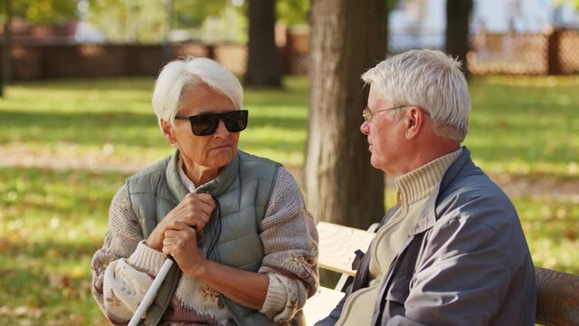 Blind elderly woman sitting on the bech with her husband. An elderly retired couple supporting each other. High quality 4k footage
