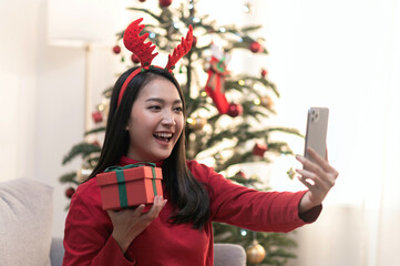 Naklejka premium Young beautiful Asian woman using smartphone connecting video call with boyfriend of selfie before open Christmas present box during Christmas festival and covid new variant omicron pandemic lockdown.