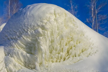 A snowdrift with icicles on the mountainside against the background of trees and blue sky in winter