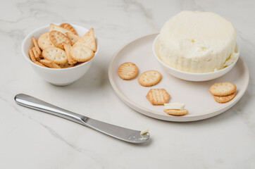 Cheese and cracker in a white bowl with a knife on a white table.