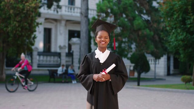 Beautiful black woman graduate posing with her diploma in front of the camera on the college garden she wearing graduation cap and suit