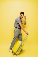 full length of cheerful young african american man adjusting sunglasses and standing with luggage on yellow.