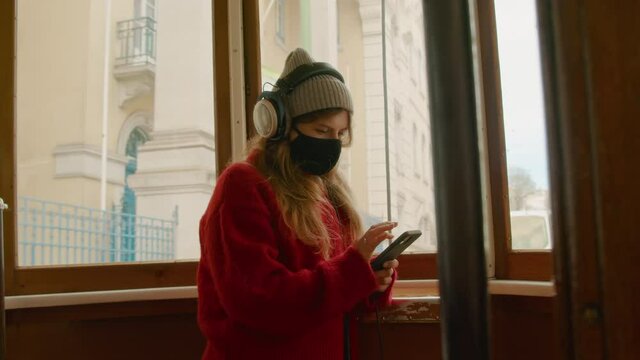 Young woman in medical covid protection face mask enjoys music in cinematic public transport. Student or tourist travel in tram in old european town, use smartphone in new normal environment