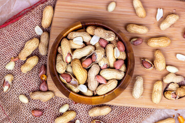 Top view of raw unpeeled peanut nuts in nutshell in brown round wooden bowl on light textile and board background.