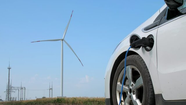 Windmill produces alternative energy for ev in field. White electric car with connected charger stands against rotating turbine near power lines