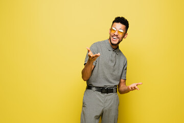 Fototapeta na wymiar positive african american man in sunglasses and grey tennis shirt gesturing isolated on yellow.