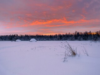 Snow-covered haystacks on the outskirts of the forest against an orange sky. Rural winter landscape