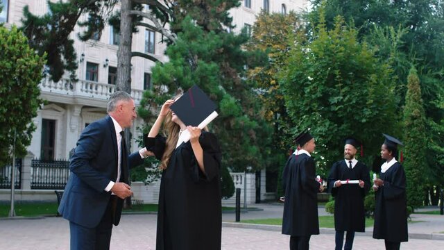 Graduation day in the college garden happy lady graduate running to her dad with diploma hugging each other and she take off the graduation cap