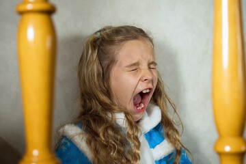 A child girl in a blue snow maiden costume yawning with her mouth wide open. The girl sits on the...