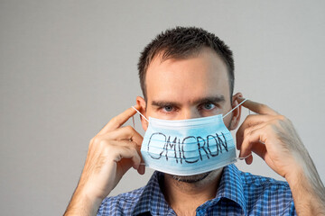 A Caucasian man in a blue plaid shirt wears a blue medical mask over his face with Omicron written...