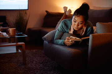 Asian woman reads book while relaxing on sofa in the evening at home.