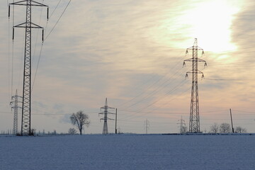 Electrical distribution network in winter landscape. High voltage pole. High voltage pylons. Infrastructure. Sun hidden in the clouds.