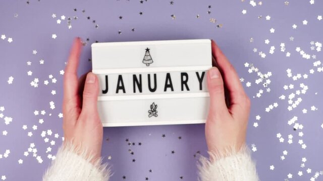 Female hands in a white fluffy sweater hold a white board with the inscription January on a purple background with glittering gold stars
