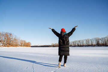 A young girl in a bright hat walks on a frozen lake. Delight and joy from the winter landscape with bright sun, clear sky. Walk in nature.
