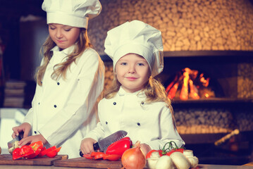 Little chefs in kitchen uniforms cut fresh vegetables with a knife.