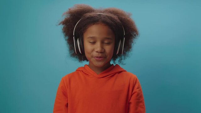 Confident African American girl in wireless headphones talking at camera standing on blue background. Child blogger speaking to camera recording video.