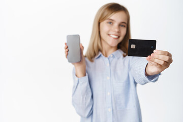 Happy blond girl, teenager smiling, showing credit card and smartphone screen, app interface, mobile banking app for children concept, standing over white background