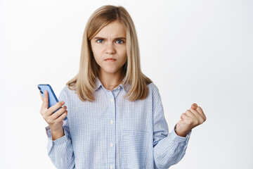 Angry little cute girl using smartphone, frowning and clenching fist threatening, boiling from anger, standing over white background