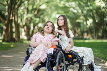 Pleasant women with disability in sitting in wheelchairs close to each other and looking in opposite sides. Female friends in summer dresses enjoying leisure time together at green park.