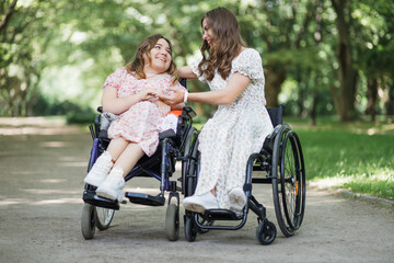 Two positive women in summer dresses smiling and looking at each other while sitting in wheelchairs among green park. Support, friendship and people concept.