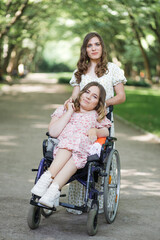 Attractive woman in dress with floral print standing at green park behind her female friend who using wheelchair. Two ladies walking together on nature. Assistance and support concept.