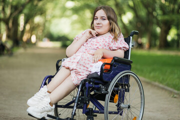 Portrait of beautiful woman with hairstyle looking at camera while sitting in wheelchair among green summer park. Lifestyles of people with chronic health disease.