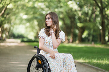 Fototapeta na wymiar Attractive caucasian woman with hairstyle looking around at green park while sitting in wheelchair. Young lady wearing stylish summer dress. Concept of people with chronic health condition.