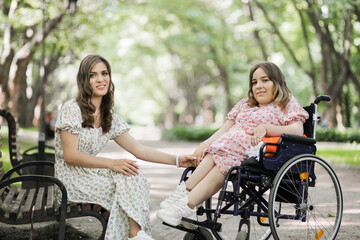 Pretty woman with disability holding hands with her female friends that sitting on wooden bench among green park. Two females enjoying common walk during summer time.