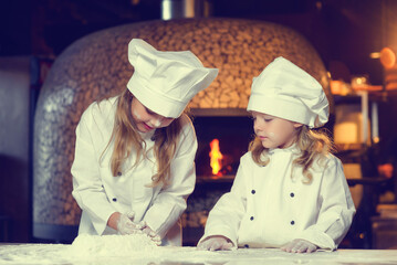 Two cheerful little girls, soiled in flour, dressed in a chef's uniform, prepare a flour product.