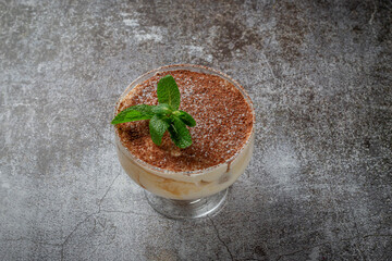 Classic tiramisu dessert with mint leaves in a glass, with cocoa and chocolate in the background of a gray stone table