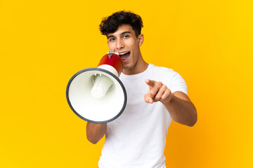 Young Argentinian man isolated on yellow background shouting through a megaphone to announce something while pointing to the front