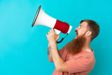 Young reddish caucasian man isolated on blue background shouting through a megaphone to announce something in lateral position