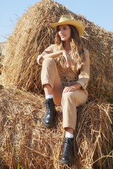 Naklejka na ściany i meble Young blonde woman in a beige jumpsuit is sitting on the rolls of hay.