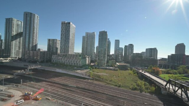 An elevated camera pans left across railway tracks and apartment towers in Toronto, Ontario, Canada. The CN Tower enters the view above the rail corridor, with pedestrian bridges under clear blue sky.