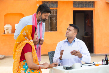 Fototapeta na wymiar Indian Doctor explaining medical test or prescribing medicine to the patient at village, woman wearing sari with her husband getting examine by medical person,Rural India healthcare concept.