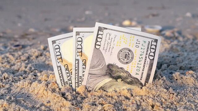 Three one hundred dollar bills half buried in sand on sandy seashore close-up. Three paper dollar bills half in sand on sea beach. Concept money, finance, investment, wealth, poverty, financing, cash
