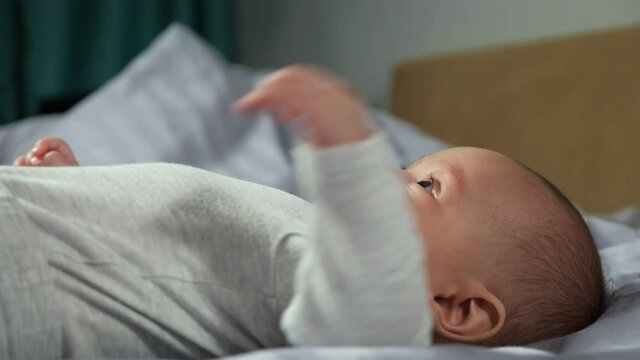 Blond-haired Baby Boy Wearing White Cotton Bodysuit Lies On Comfortable Blanket And Moves Hands Blowing Bubbles From Saliva, Closeup.