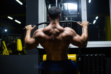 Fototapeta na wymiar Handsome fitness man is performing back workouts using thrust of the upper block machine in a gym, back view
