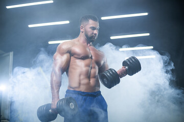 Front view of a muscular bodybuilder lifting dumbbells in blue shorts posing in a gym standing in...