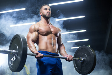 Fototapeta na wymiar Bottom up view of a muscular athlete lifting heavy barbell showing his muscles in a gym standing in smoke