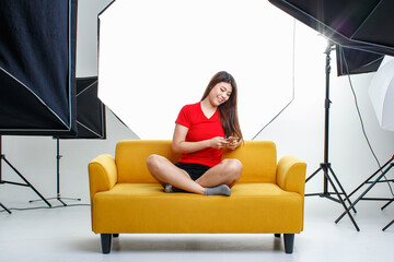 Portrait studio shot of Asian pretty thoughtful female model in red shirt sitting thinking on cozy yellow sofa couch on photographing shooting backstage set with softbox lighting reflector spotlight