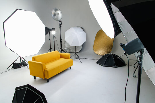 Studio shot fashion backstage photographing shooting set with yellow cozy sofa couch and photography equipment softbox flash strobe spotlight tripods reflector umbrella on white backdrop background