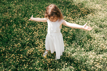 Naklejka na ściany i meble Little girl in white dress, arms outstretched, stands in middle of blooming meadow. Life insurance. Carefree childhood. Summer days. Beauty of nature. Walks and adventures