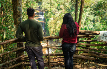 young couple watching waterfall falling from mountain top in green forests at morning from top angle