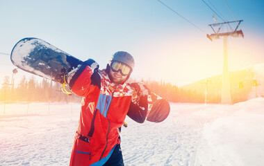 Portrait of young bearded man with helmet holding snowboard against backdrop of ski resort and lift sunlight. Concept winter sport banner