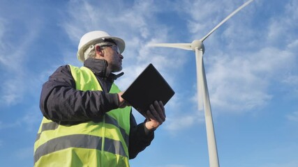 Low angle shot of renewable energy technician operator using tablet,wind turbine power plant background,engineer worker checking wind turbine does maintenance to power station generator